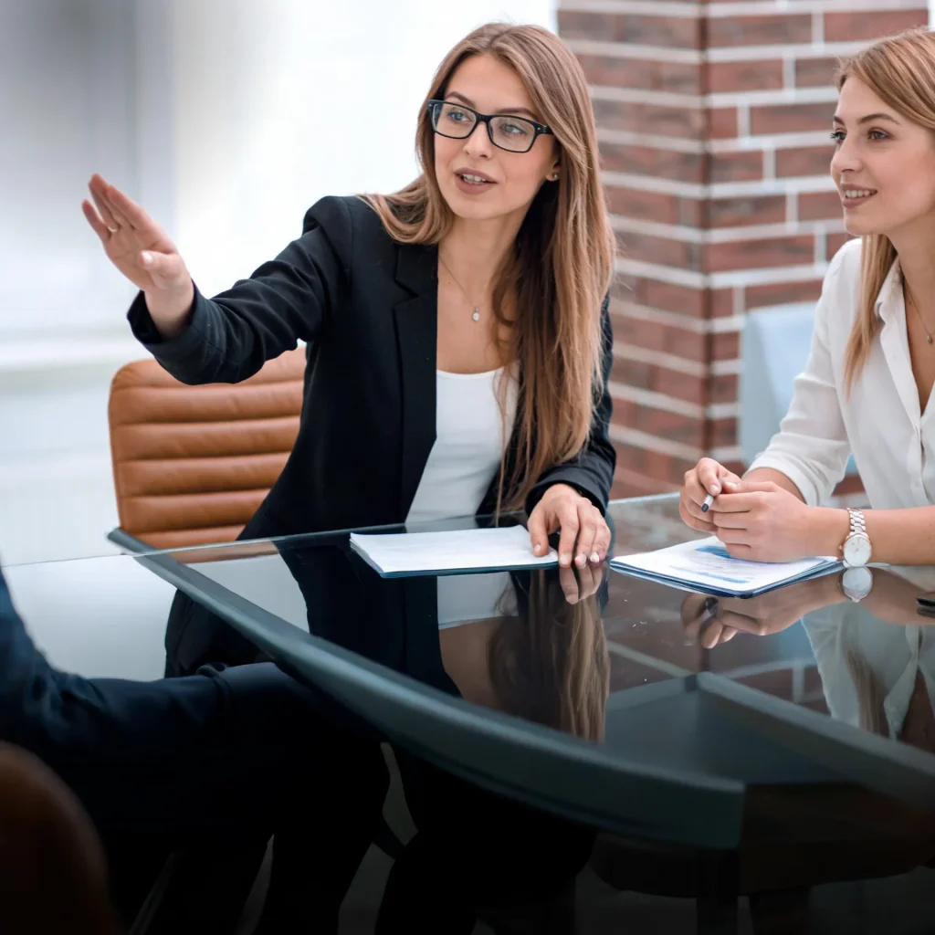 Two businesswomen sit at a glass conference table the woman in a black blazer gestures as she talks to her colleague next to her