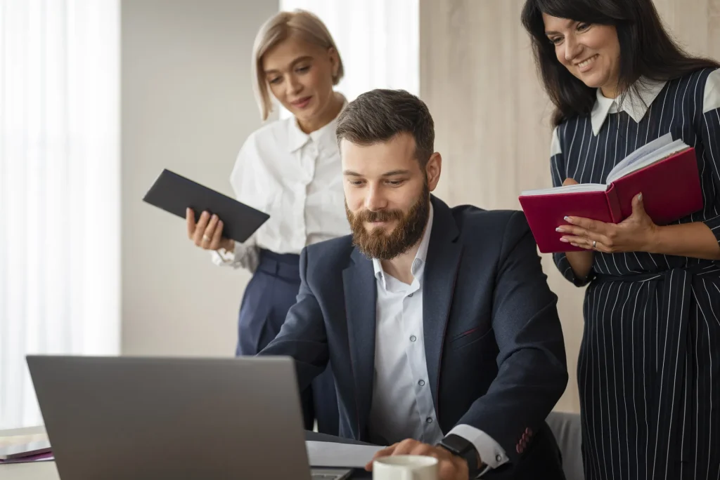 Bearded man in a suit typing on a laptop as two female colleagues stand beside him one holding a black folder and the other a red notebook smiling in an office